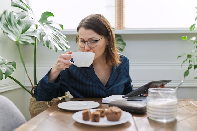 Grave mujer madura que trabaja en casa. Mujer en pijama sentada a la mesa por la mañana, desayunando y usando tableta digital, cuaderno de negocios