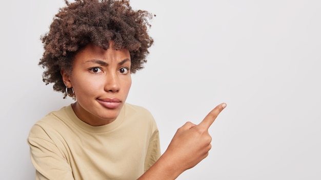 Grave mujer de pelo rizado muestra puntos algo desagradables con el dedo en la pared en blanco muestra que el anuncio del producto recomienda probar el artículo suscribirse o hacer clic en el enlace aislado sobre fondo blanco