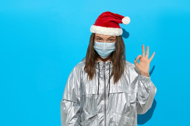 Grave mujer vistiendo gorro de Papá Noel y máscara protectora, mostrando gesto OK