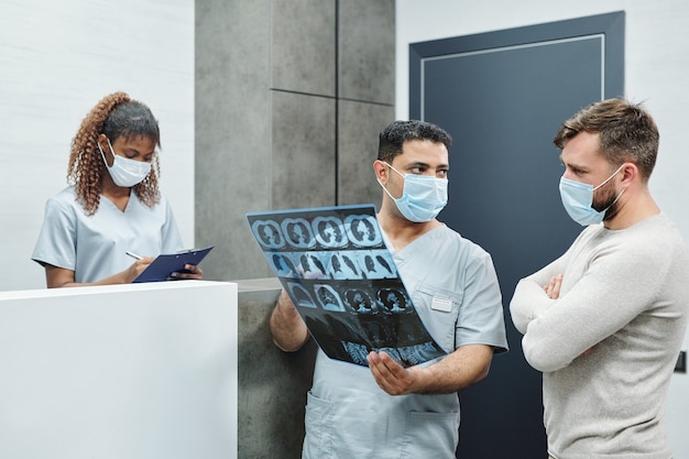 Foto grave radiólogo masculino de raza mixta en uniforme y máscara que muestra la imagen de rayos x al paciente barbudo contra el mostrador de recepción y la joven enfermera