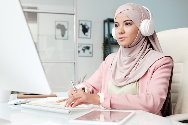 Grave religiosa joven musulmana en auriculares sentado en el escritorio de oficina y trabajando con la computadora