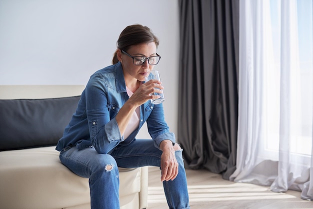 Grave triste mujer madura de mediana edad sentada en su casa sosteniendo un vaso de agua mirando por la ventana. Problemas de salud, dificultades mentales, depresión, menopausia y problemas personales.