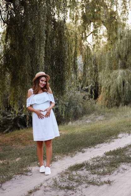 grávida bonita abraça sua barriga. caminhar no parque. garota em um vestido branco