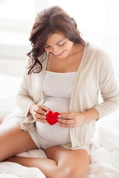 gravidez, amor, pessoas e conceito de expectativa - mulher grávida feliz com coração vermelho na cama no quarto de casa