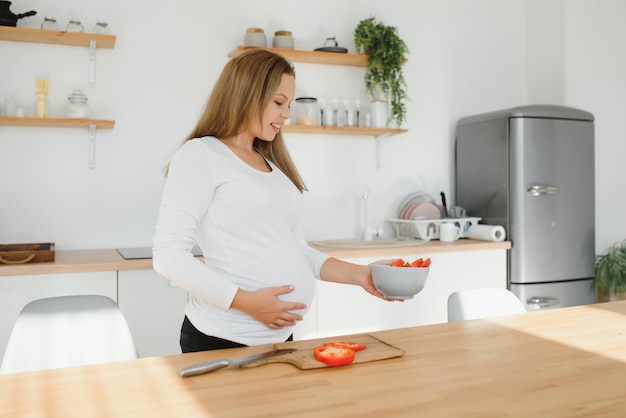 Gravidez, cozinhar alimentos, estilo de vida saudável, pessoas e conceito de expectativa - mulher grávida feliz com uma faca cortando pepino e preparando salada de legumes em casa