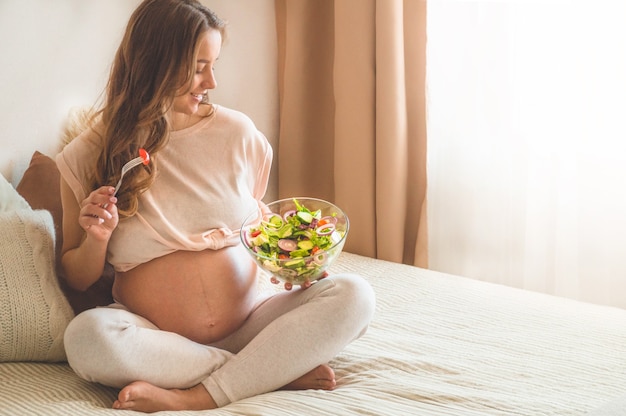 Gravidez e nutrição orgânica saudável. Mulher grávida comendo salada de legumes fresca na cama, espaço livre