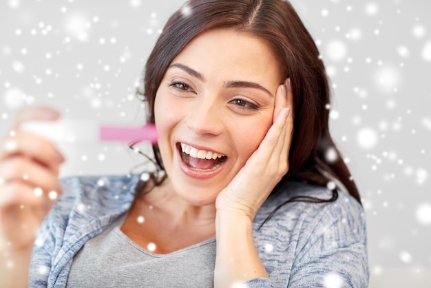 Foto gravidez, fertilidade, maternidade, emoções e conceito de pessoas - mulher sorridente feliz olhando para o teste de gravidez em casa sobre a neve