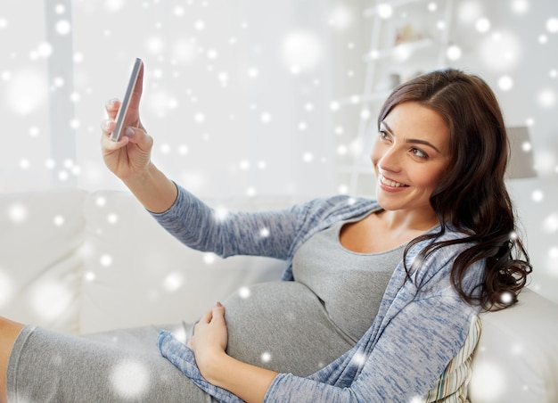 Foto gravidez, inverno, tecnologia, pessoas e conceito de expectativa - mulher grávida feliz com smartphone tirando selfie em casa sobre a neve