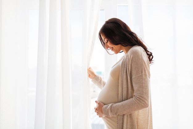 gravidez, maternidade, pessoas e conceito de expectativa - close-up de uma mulher grávida feliz com barriga grande na janela
