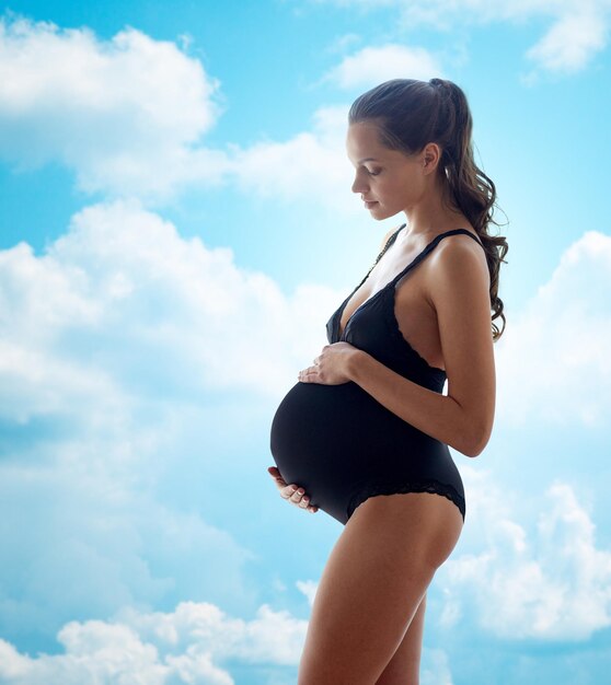 Gravidez, maternidade, pessoas e conceito de expectativa - mulher grávida feliz em cueca preta sobre fundo de céu azul e nuvens