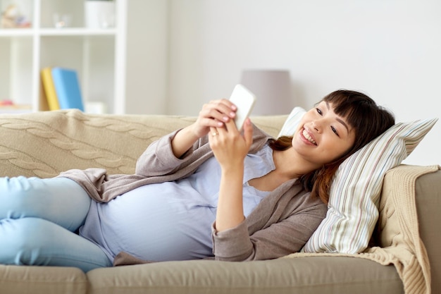 gravidez, maternidade, tecnologia e conceito de pessoas - mulher asiática grávida feliz com smartphone deitado no sofá em casa