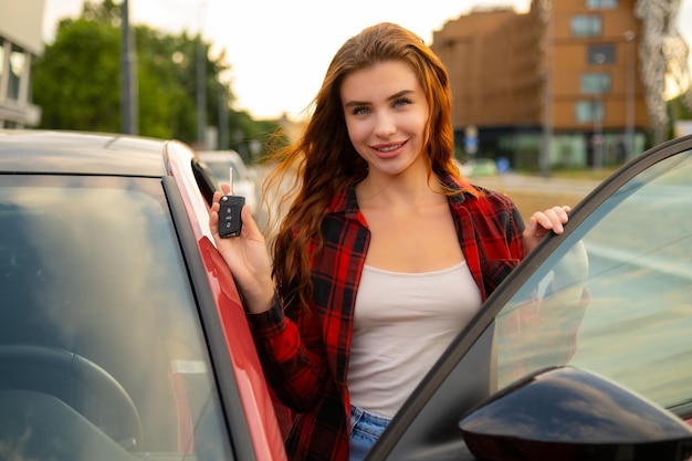 Graziös neben einem kürzlich gekauften roten Auto steht eine rothaarige Frau mit den Schlüssel