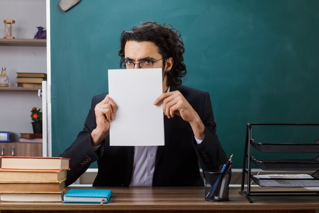 Greddy männlicher Lehrer mit Brille hält und bedecktes Gesicht mit Papier sitzt am Tisch mit Schulwerkzeugen im Klassenzimmer