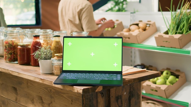 Greenscreen-Laptop im örtlichen Öko-Laden mit Kisten mit organischen heimischen Erzeugnissen und Bulk-Produkten in Gläsern auf Regalen PC mit isolierter Anzeige mit Mockup-Copyspace-Vorlage