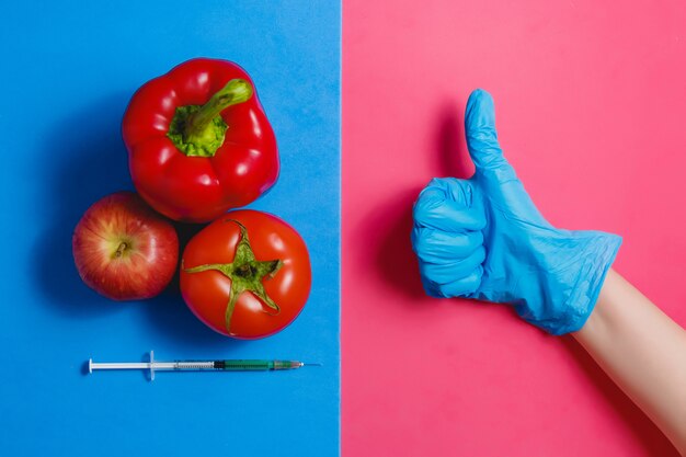 Greifen Sie herauf grüne Flüssigkeit in der Spritze, rote Tomate, Apfel, Pfeffer ab. Genetisch geändertes Nahrungsmittelkonzept auf rosafarbenem Blau.