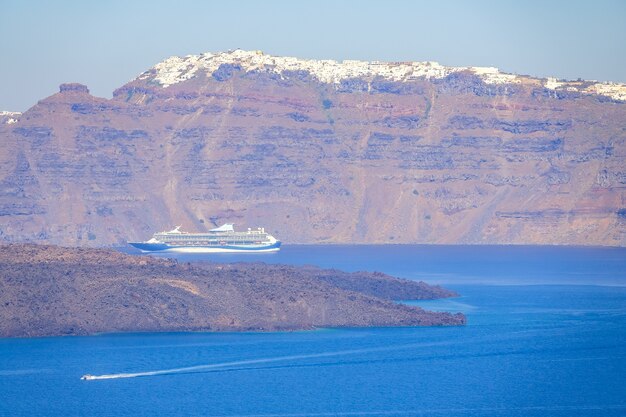 Griechenland. Sonniger Sommertag vor der felsigen Küste von Santorin. Großes Kreuzfahrtschiff und kleines Motorboot