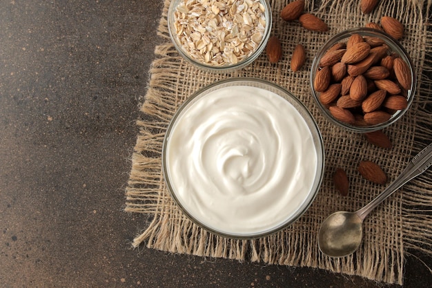 Griechischer Joghurt in einer Glasschüssel neben einem Löffel Haferflocken und Mandeln auf dunklem Hintergrund Gesundes Essen Naturjoghurt Frühstück Draufsicht mit Platz für Text