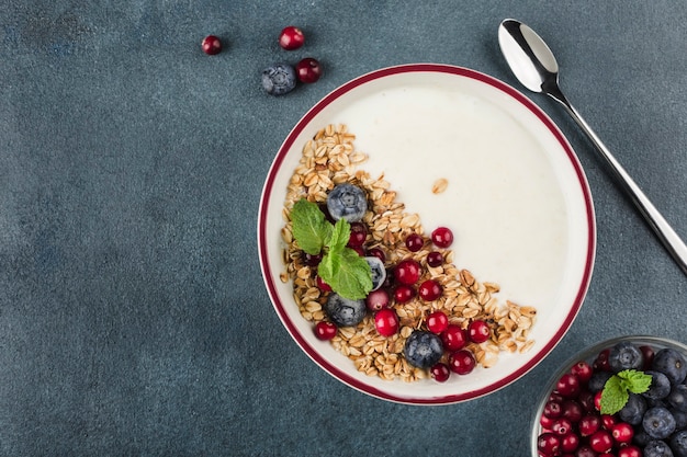 Griechischer Joghurt mit Müsli, frischen Blaubeeren, Preiselbeeren und Minze in einem Teller mit einem Löffel auf dunkelblauem Hintergrund. Ein leichtes Frühstück für Diät und Fitness. Draufsicht mit einem Kopienraum für den Text.