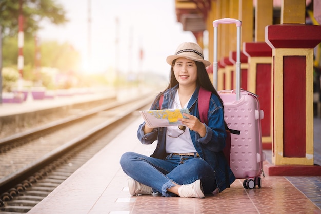 Gril asiático joven que sostiene el mapa y que se sienta en la estación de tren antes de viaje. Concepto de trabajo y viaje.