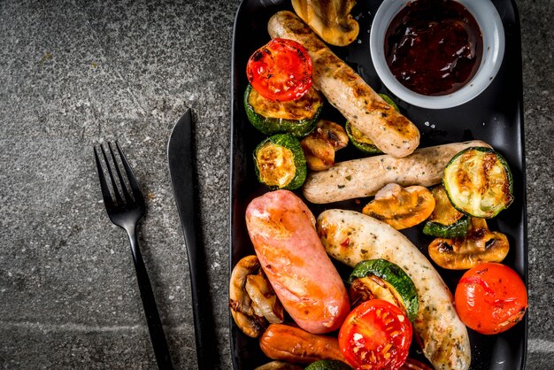 Grill. Auswahl an verschiedenen gegrillten Fleischwürsten, mit Gemüse BBQ - Pilze, Tomaten, Zucchini, Zwiebeln. Auf einem schwarzen Steintisch, auf einem schwarzen Teller, mit Soße.