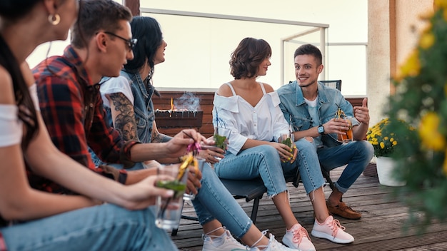 Grillen Sie junge, fröhliche Leute, die sich unterhalten und Cocktails trinken, während sie auf dem Dach sitzen