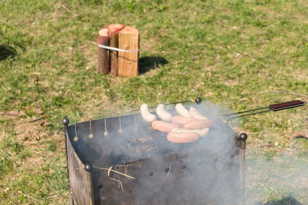 Grillen von leckeren Fleischwürsten im Freien zum Mittagessen auf einem Barbecue-Grill im Park an einem sehr sonnigen Tag.