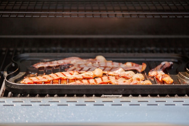 Grillen von Speckstreifen auf einer gusseisernen Grillplatte im Gasgrill im Freien.