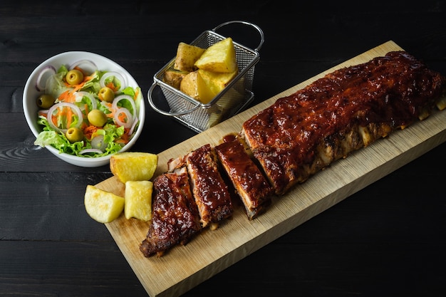 Grillrippen mit Kartoffeln und Salat auf einem Holzbrett auf einem dunklen Hintergrund. Speicherplatz kopieren.