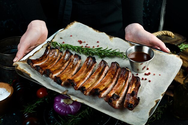 Grilltablett mit gebackenen saftigen Schweinerippchen in den Händen des Küchenchefs auf rustikalem Holzhintergrund