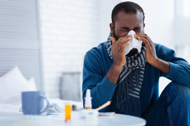 Gripe insuportável. Belo jovem doente sentado em seu quarto confortável e espirrando