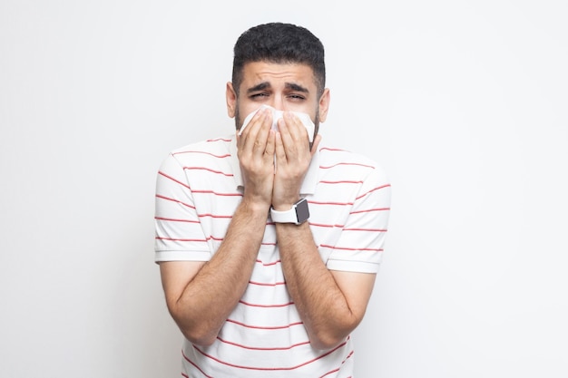 Gripes e resfriados. Retrato de um jovem barbudo doente em pé de camiseta listrada, segurando o tecido na boca e olhando para a câmera com tristeza e doença. tiro do estúdio interno, isolado no fundo branco.