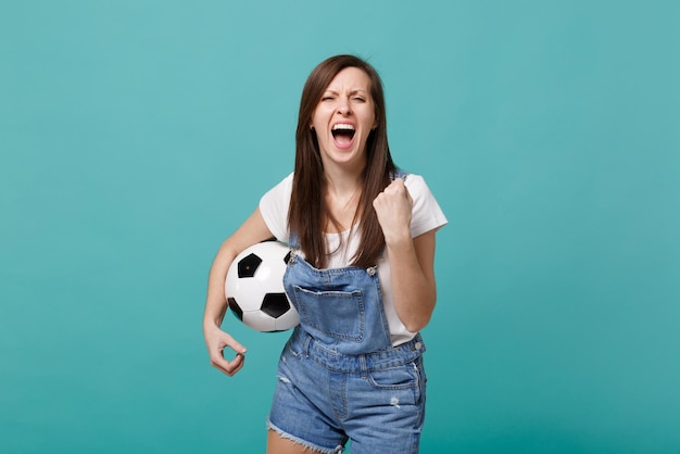 Gritando loca joven hincha de fútbol animar apoyo equipo favorito con balón de fútbol haciendo gesto de ganador aislado sobre fondo azul turquesa de la pared. Emociones de la gente, concepto de ocio familiar deportivo.