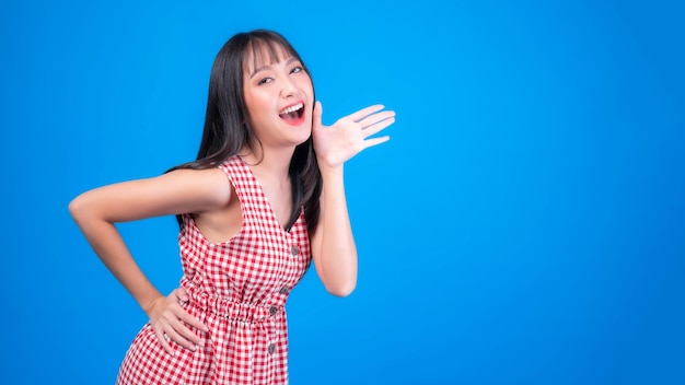 Gritos emocionados, hermosa mujer asiática linda chica en vestido rojo con flequillo gritos de peinado anuncian las buenas noticias o promoción, tomados de la mano cerca de su cara con la boca abierta promoción de noticias Herald