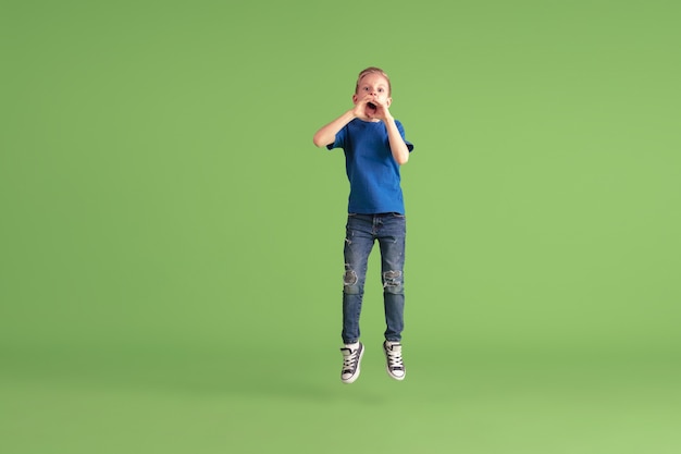 Gritos. Niño feliz jugando y divirtiéndose en la pared verde. Niño caucásico en tela brillante se ve juguetón, riendo, sonriendo. Concepto de educación, infancia, emociones, expresión facial.