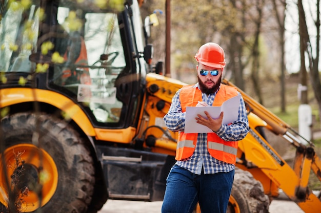 Grober Bartarbeitskraftmann-Klagenbauarbeiter im orange Sturzhelm der Sicherheit, Sonnenbrille gegen traktor mit Planpapier an den Händen.