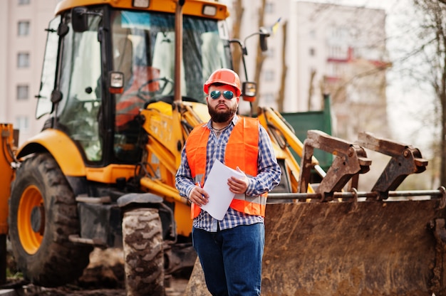 Grober Bartarbeitskraftmann-Klagenbauarbeiter im orange Sturzhelm der Sicherheit, Sonnenbrille gegen traktor mit Planpapier an den Händen.