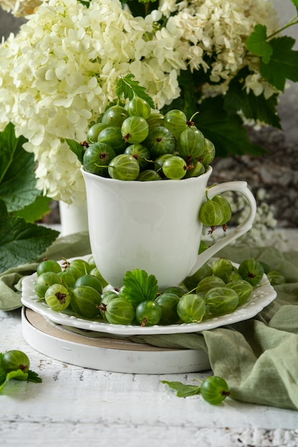 Grosellas espinosas en plato y taza sobre la mesa. Frutas maduras de verano y flores en florero composición botánica