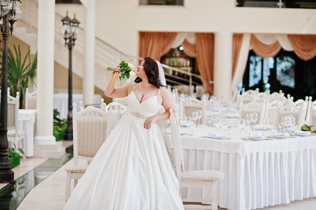 Große Brüste Brunettebraut mit dem Hochzeitsblumenstrauß warfen an den Hochzeitssaal-Hintergrundtabellen auf