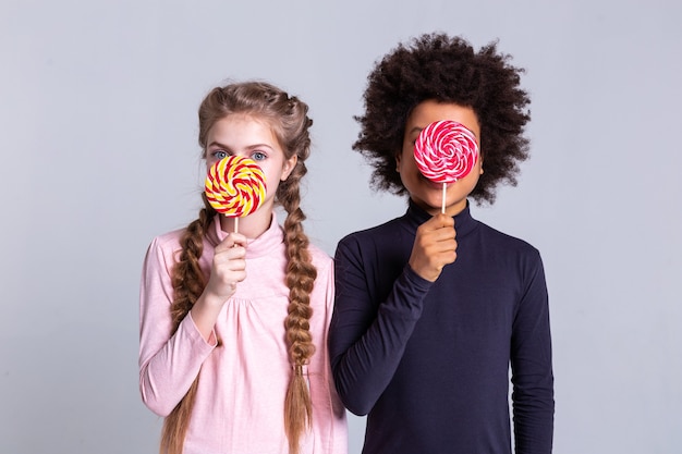 Große bunte Bonbons. Hübsches kleines Mädchen mit großen blauen Augen und langen hellen Zöpfen, die ihr Gesicht mit einem lockigen Jungen bedecken