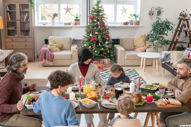 Große Familie bestehend aus drei Generationen, die an einem festlich servierten Tisch sitzen, sich unterhalten und hausgemachtes Essen zum Weihnachtsessen im Wohnzimmer genießen