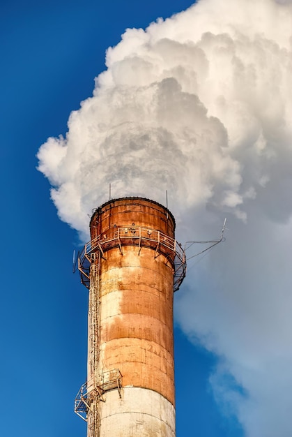 Große grungige Fabrikpfeife, die dicken weißen Rauch gegen wolkenlosen blauen Himmel ausstrahlt