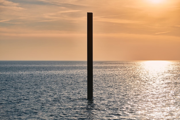 Große rostige Stahlkonstruktionssäule im blauen Meer mit ruhigen Wellen bei Sonnenuntergang, Sonnenlicht reflektiert auf der Meerwasseroberfläche. Vertikales verlassenes Gebäudeteil, Baustelle nahe Küste