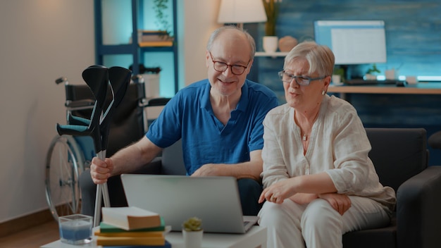 Großeltern winken der Webcam für Videokonferenzen zu und chatten mit Laptop. Mann mit Krücken und Frau sprechen über Online-Ferntelefonkonferenz. Alte Leute, die sich im Internet unterhalten