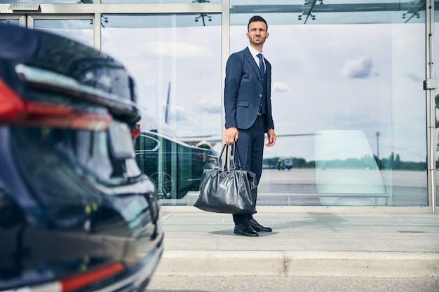 Großer, gutaussehender, brünetter Mann, der eine Tasche hält, während er vor einem Terminal steht und auf den Transport wartet