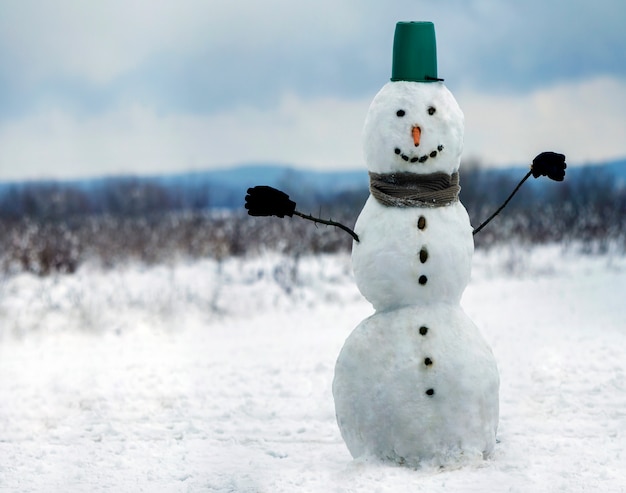 Großer lächelnder Schneemann mit Eimerhut, Schal und Handschuhen auf weißer Schneefeldwinterlandschaft, verschwommenen schwarzen Bäumen und blauem Himmel kopieren Raumhintergrund. . Frohe Weihnachten und ein frohes neues Jahr Grußkarte.