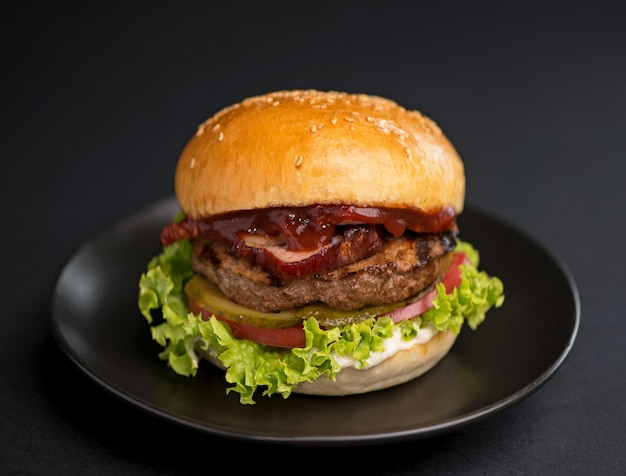 Großer leckerer Burger mit Rindfleisch-Patty-Salat-Speck und Essiggurke auf schwarzem Hintergrund