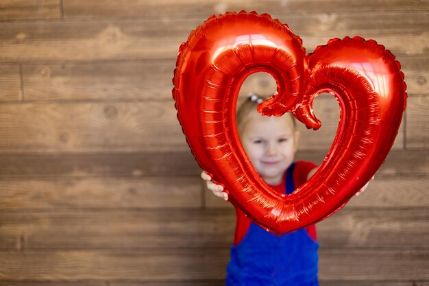 Großes Ballonherz im Fokus in den Händen eines glücklichen kleinen Mädchens auf einem Holzwandhintergrund Valentinstag Konzept leerer Raum für Text