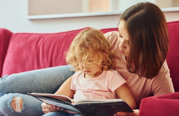 Großes Buch lesen Junge Mutter mit ihrer kleinen Tochter in Freizeitkleidung zusammen zu Hause