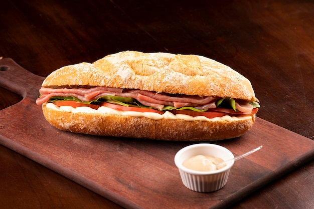 Großes geräuchertes Carpaccio-Sandwich mit Rucola-Tomaten und Gruiere-Käse auf italienischem Baguette auf Holzbrett und dunkler Hintergrundwinkelansicht mit Mayonnaise von vorne