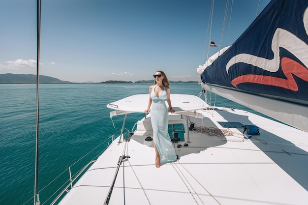 Großes, langbeiniges, sexy junges Model, das ein langes blaues Kleid und eine Sonnenbrille trägt und auf ihrer weißen Laxuryacht vor dem Hintergrund von Wasser und Natur posiert. Urlaubskonzept